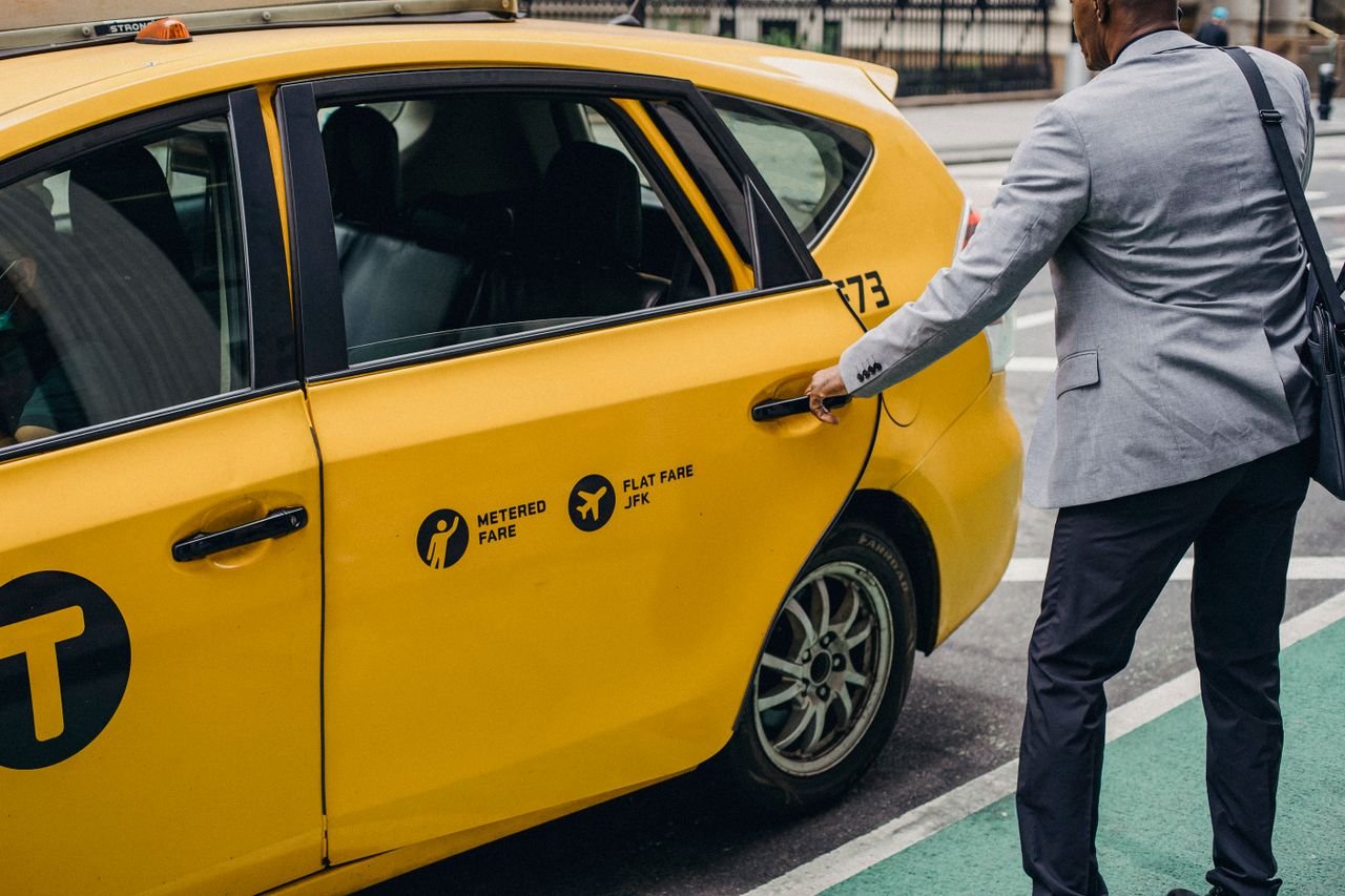 Back view of crop unrecognizable African American male entrepreneur in formal clothes opening door of yellow taxi parked on city street